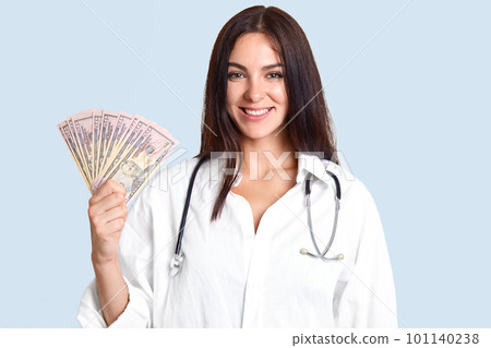 Medicine and corruption concept. Happy young professional female doctor holds money in hands, happy to recieve bribe from patients, wears white robe and phonendscope, isolated over blue background 101140238