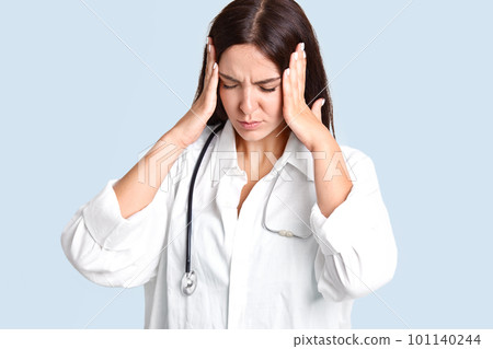 Stressful female family doctor suffers from headache, keeps palms on temples, closes eyes, has pressure on work, feel fatigue after work all day in clinic or hospital, poses indoor. Healthcare concept 101140244