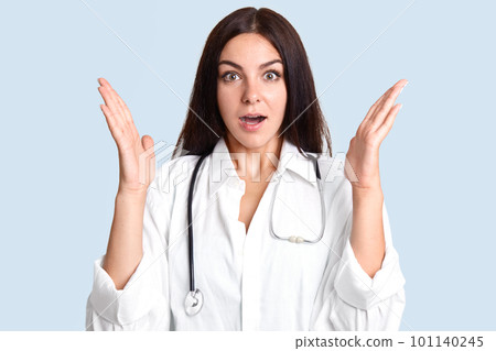 Shocked young female therapist clasps hands and looks surprisingly at camera, can`t believe in death of patient, has scared look, dressed in lab gown, gestures indoor against blue background 101140245