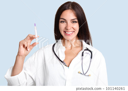 Cheerful brunette female doctor with stethoscope and syringe going to give medical treatment to patient, has friendly look, isolated over blue background. Woman general practitioner in hospital Cheerful brunette female doctor with stethoscope and syringe going to give medical treatment to patient, has friendly look, isolated over blue background. Woman general practitioner in hospital 101140261