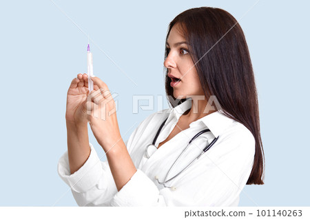Sideways shot of female physician holds syringe, gong to make vaccination, looks at remedy with shocked expression, poses against light blue background, being professional specialist in medical sphere Sideways shot of female physician holds syringe, gong to make vaccination, looks at remedy with shocked expression, poses against light blue background, being professional specialist in medical sphere 101140263