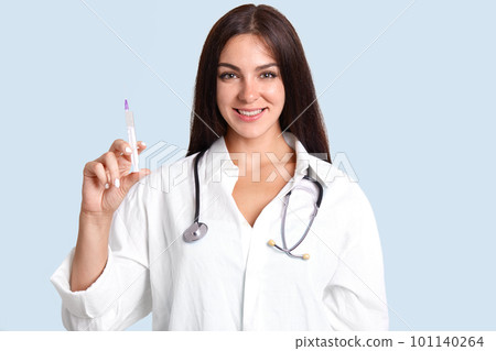 Beautiful young female doctor in white robe, has phonendoscope on neck, holds syringe with liquid, isolated over blue background, has cheerful expression. Glad woman nurse makes injections to patient Beautiful young female doctor in white robe, has phonendoscope on neck, holds syringe with liquid, isolated over blue background, has cheerful expression. Glad woman nurse makes injections to patient 101140264