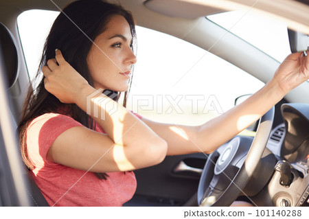 Shot of beautiful brunette woman dressed in casual t shirt, looks seriously at mirror of car, has thoughtful expression, concenrated into distance. Horizontal view of gorgeous lady in automobile 101140288