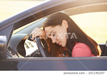 Transportation concept. Tired beautiful woman driver has sleepy expression, stop on road as can`t drive any more, wants to have rest, leans at wheel, poses in car, feels overworked and fatigue Transportation concept. Tired beautiful woman driver has sleepy expression, stop on road as can`t drive any more, wants to have rest, leans at wheel, poses in car, feels overworked and fatigue 101140297