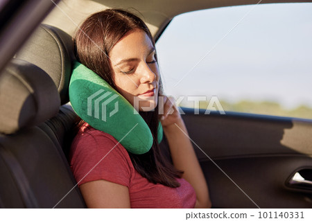 Fatigue brunette woman uses green pillow for travelling in car, has tired look, dressed in casual t shirt, poses in automobile, falls asleep. People, tourism and journey concept. Female enjoys comfort 101140331