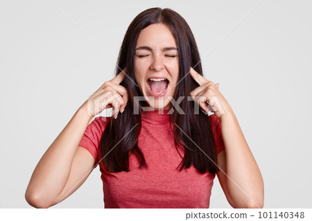 Annoyed brunette woman asks to stop this sound, pluggs ears with both index fingers, exclaims angrily, keeps eyes shut, dressed casually, isolated over white background. Aggressive girl ignores sound Annoyed brunette woman asks to stop this sound, pluggs ears with both index fingers, exclaims angrily, keeps eyes shut, dressed casually, isolated over white background. Aggressive girl ignores sound 101140348