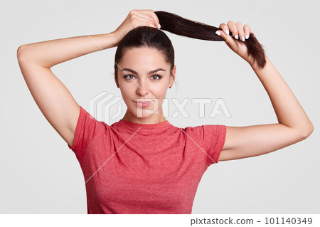 Beautiful brunette female touches pony tail, has healthy skin, dressed in casual t shirt, has make up, rests at home, shows her natural beauty isolated over white background. Young woman models indoor Beautiful brunette female touches pony tail, has healthy skin, dressed in casual t shirt, has make up, rests at home, shows her natural beauty isolated over white background. Young woman models indoor 101140349