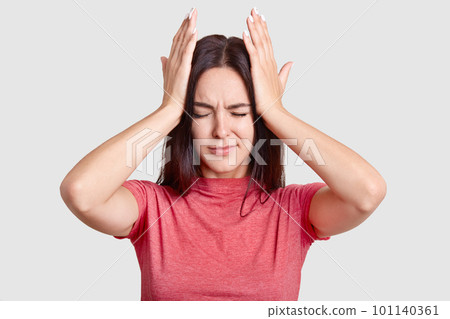 Photo of brunette woman with desperate look, keeps both hands on head, suffers from terrible headache, dressed in casual t shirt, poses against white background. People and tiredness conncept 101140361