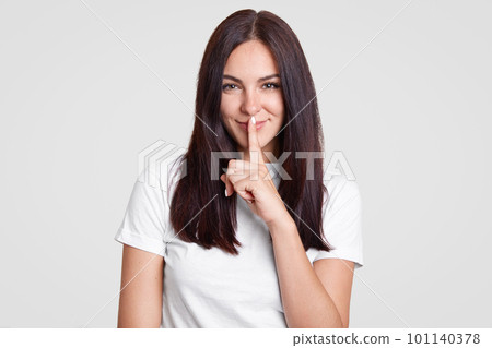 Attractive woman with long dark hair has secret, keeps fore finger over mouth, shows shush gesture, has satisfied facial expression, dressed in casual outfit, isolated over white background. Attractive woman with long dark hair has secret, keeps fore finger over mouth, shows shush gesture, has satisfied facial expression, dressed in casual outfit, isolated over white background. 101140378
