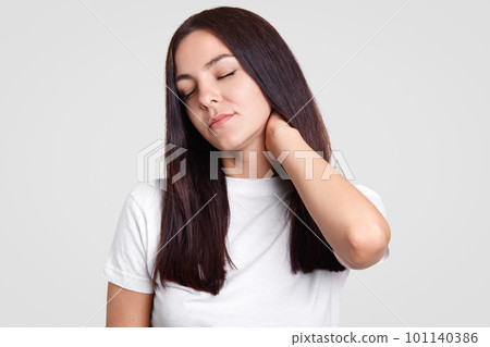 Tired beautiful brunette female keeps hand on neck, feels pain, tired after working in office, dressed in casual white t shirt, isolated over studio wall. Fatigue woman feels sick. Overworking concept 101140386