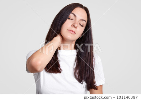 Photo of tired brunette female feels pain in neck as has sedentary lifestyle, needs physical activity, closes eyes, wants sleep, have rest, dressed in casual white t shirt, stands against studio wall 101140387