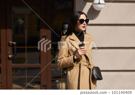 Beautiful brunette young woman holds cup of coffee in her hands, walks along sunlighted streets, wears long fashionable mustard coat, has black accessories, bright makeup. Street fashion concept. 101140409