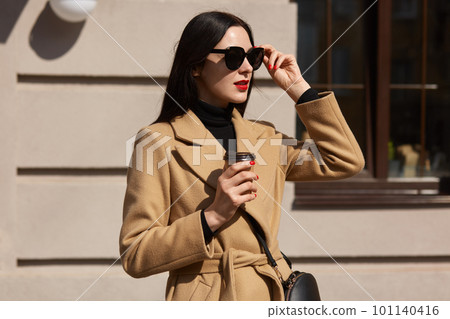 Outdoor fashion photo of young beautiful isolated over street building background, elegant lady lifting her sunglasses and looking away, enjoys drinking hot takeaway beverage while walking alone. 101140416