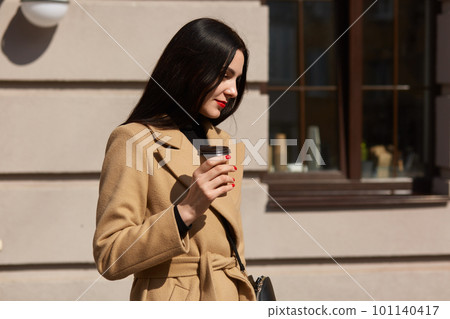 Young beautiful woman in beige coat with hand bag walking on street isdolated over cafe background, has straight long dark hair, has bright red manicure, businesslady after meeting. Business concept. 101140417