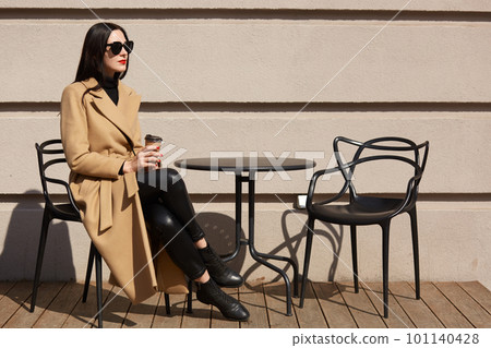 Shot of woman wearing fashionable coat having breakfast in cozy street outdoor cafe and drinking coffee from paper mug. Girl sits at restaurant terrace, thinking or dreaming about something important. 101140428