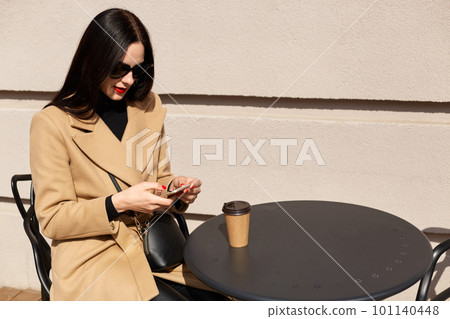 Close up portrait of beautiful elegant young woman wearing beige coat and drinking takeaway coffee while having lunch time. Pretty brunette lady checking her social networking sites, chatting. 101140448