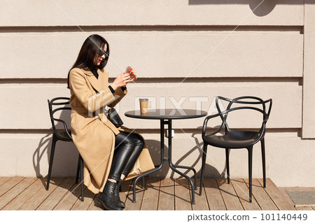 Slender black haired female sits at table outdoors, having cup of hot drink, relaxing during free time, taking photos of urban scenes in warm sunny spring daytime. People and urban space concept. 101140449