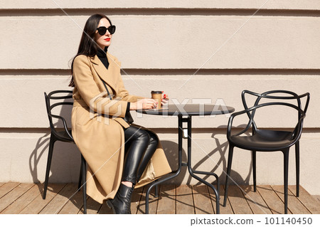 Pleased beautiful young model poses isolated sitting over light wall on terrace, having coffee break, enjoys sunny day, looking aside. Slender pretty female wearing stylish beige coat and black pants. 101140450