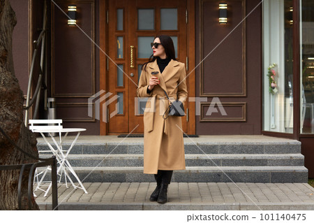 Beautiful young brunettee woman in modern beige coat, holding disposable takeaway cup and standing against urban city building, looks thoughtfuly aside, has dating, waiting for her boyfriend. 101140475