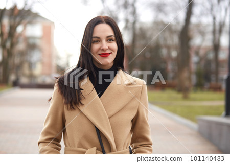 Half length shot of attractive young woman in elegant beige coat walking on street. Adorable brunette woman spends her leisure time in park, has pleasant facial expression, being in good mood. Half length shot of attractive young woman in elegant beige coat walking on street. Adorable brunette woman spends her leisure time in park, has pleasant facial expression, being in good mood. 101140483