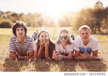 Glad four friends with happy expression lie on grass, hold chins and look positvely at camera, enjoy sunshine, have good rest, like to spend free time outdoor. Youth, teenagers and lifestyle concept Glad four friends with happy expression lie on grass, hold chins and look positvely at camera, enjoy sunshine, have good rest, like to spend free time outdoor. Youth, teenagers and lifestyle concept 101140593