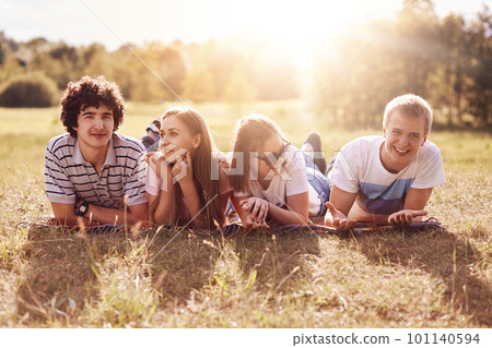 Horizontal shot of four happy friends lie on ground, have picnic during sunny summer day, have positive expressions, smile pleasantly at camera. Funny companions pleased to spend leisure time together Horizontal shot of four happy friends lie on ground, have picnic during sunny summer day, have positive expressions, smile pleasantly at camera. Funny companions pleased to spend leisure time together 101140594