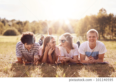 Two female fellows have pleasant talk while lie on stomach on green grass near their boyfriends, enjoys sunny summer time all together, have picnic during weekend. Friends and recreation concept Two female fellows have pleasant talk while lie on stomach on green grass near their boyfriends, enjoys sunny summer time all together, have picnic during weekend. Friends and recreation concept 101140595