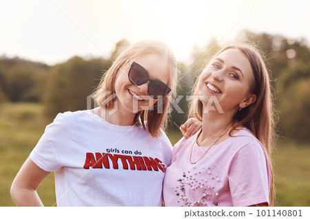 Two positive female friends wear t shirt and shades, spend free time on nature, smile pleasantly at camera, have good relationship. Positive teenagers have summer vacation outside. Leisure concept 101140801