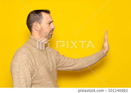 Rejection, denial concept. Side view of man wearing turtleneck making stop sign with outstretched hand, warning expression with negative gesture. Indoor studio shot isolated on yellow background 101140862