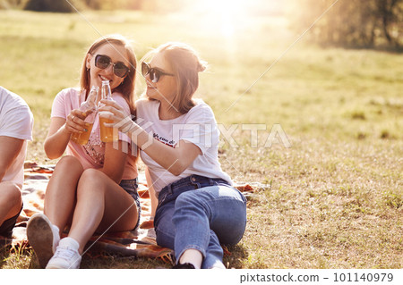 Cheerful young female companions clink bottles of energy drink, have fun together outdoor, wear trendy sunglasses, casual clothes, spend leisure time on nature, have picnic and unforgettable day 101140979