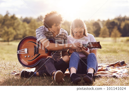 Picnic time. Lovely couple in love date on nature background, handsome male with curly hair teaches his beautiful girlfriend to play guitar, have good relationship. Dating, romance, lifestyle concept Picnic time. Lovely couple in love date on nature background, handsome male with curly hair teaches his beautiful girlfriend to play guitar, have good relationship. Dating, romance, lifestyle concept 101141024