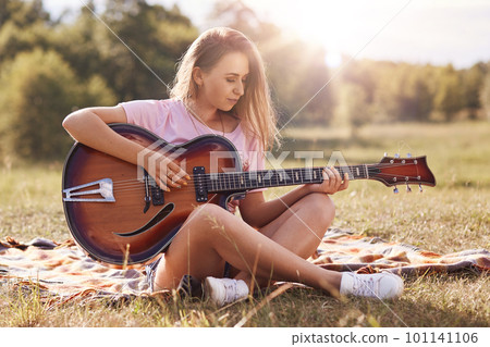 Beautiful young woman playing gitar in meadow, has stright blonde hair, looks concentrated on musical insrtument, wearing casual clothes, spending time alone, enjoying wonderful nature. Outdoor shot 101141106