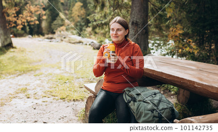 Happy redhead Woman in active trekking clothes having a halt after hiking. Hiker drinking water from water bottle or hot drink from yellow thermos Happy redhead Woman in active trekking clothes having a halt after hiking. Hiker drinking water from water bottle or hot drink from yellow thermos 101142735