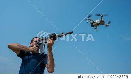 A man aims to shoot a rifle at a flying drone against a blue sky.  101142739