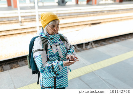 Old happy woman with backpack in yellow hat waiting train on station platform and using smart phone on urban background. Railroad transport concept 101142768