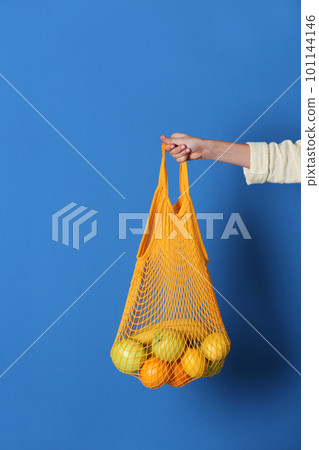 Woman holding net bag with fruits on blue background, closeup Woman holding net bag with fruits on blue background, closeup 101144146