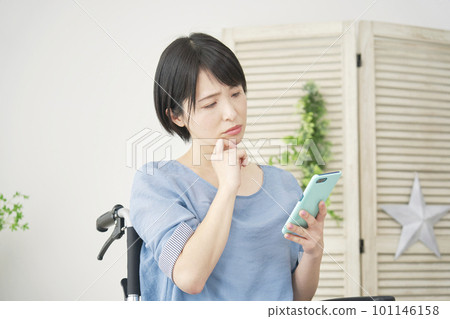 A woman on a wheelchair thinking while using a smartphone in the living room A woman on a wheelchair thinking while using a smartphone in the living room 101146158
