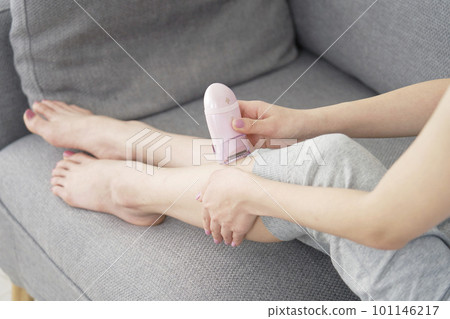 A woman doing self hair removal on her legs with a home epilator in the living room 101146217