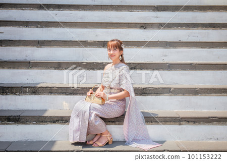Young woman wearing traditional Thai dress sits holding an antique bag at the Wat Arun Temple, a popular destination for tourists around the world. Bangkok Thailand Young woman wearing traditional Thai dress sits holding an antique bag at the Wat Arun Temple, a popular destination for tourists around the world. Bangkok Thailand 101153222