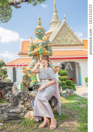 Young woman wearing traditional Thai dress with accessories sitting in front of a giant at Wat Arun Ratchawararam The most popular destination for tourists around the world. Bangkok, Thailand Young woman wearing traditional Thai dress with accessories sitting in front of a giant at Wat Arun Ratchawararam The most popular destination for tourists around the world. Bangkok, Thailand 101153225