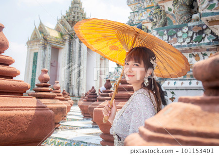 Young woman wearing traditional Thai dress stands holding an ancient umbrella at Wat Arun, a popular destination for tourists around the world. Bangkok Thailand Young woman wearing traditional Thai dress stands holding an ancient umbrella at Wat Arun, a popular destination for tourists around the world. Bangkok Thailand 101153401