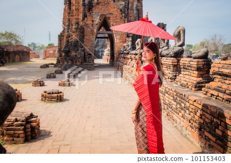 young woman wearing traditional red Thai dress and golden accessories stands holding a traditional umbrella in the historical site Wat Chaiwatthanaram Ayutthaya. Thai national costume young woman wearing traditional red Thai dress and golden accessories stands holding a traditional umbrella in the historical site Wat Chaiwatthanaram Ayutthaya. Thai national costume 101153403