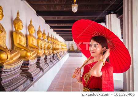 Young woman wearing traditional red Thai dress and golden accessories stands holding a traditional umbrella in the historical site Wat Phutthaisawan Ayutthaya. Thai national costume Young woman wearing traditional red Thai dress and golden accessories stands holding a traditional umbrella in the historical site Wat Phutthaisawan Ayutthaya. Thai national costume 101153406