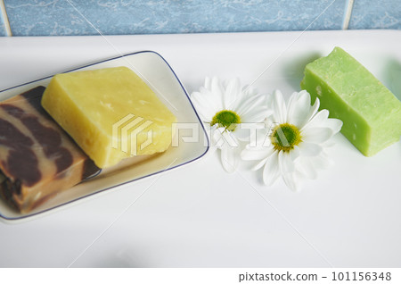 Selective focus on chamomile flowers and hand-made soap bars with natural herbal ingredients on white ceramic wash basin. Organic herbal cosmetic products for home spa, beauty treatment and body care 101156348
