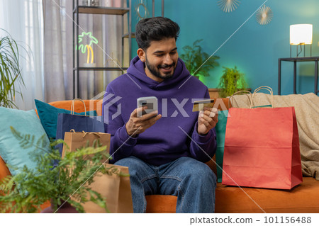 Happy shopaholic indian man sitting with shopping bags making online payment with credit card 101156488