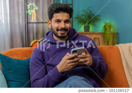 Indian hindu young man sitting on sofa, using smartphone share messages on social media application Indian hindu young man sitting on sofa, using smartphone share messages on social media application 101156517