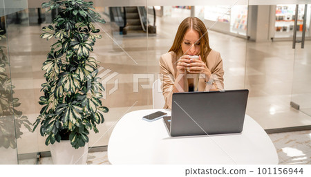 A business woman sits in a cafe, works at a computer, drinks coffee. She is wearing a beige jacket, brown top and black trousers. A business woman sits in a cafe, works at a computer, drinks coffee. She is wearing a beige jacket, brown top and black trousers. 101156944