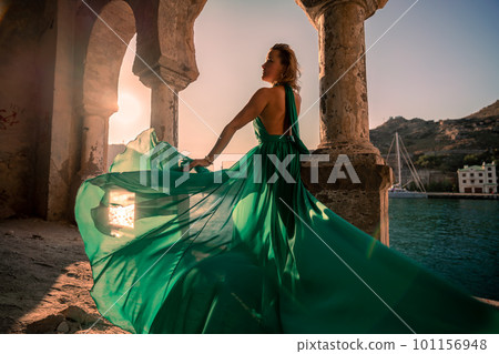 Rear view of a happy blonde woman in a long mint dress posing against the backdrop of the sea in an old building with columns. Girl in nature against the blue sky. 101156948
