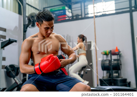 Asian sport man sit on chair and try to wear boxing glove in front of the woman exercise in the back 101164401