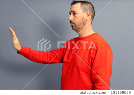 Stop. Side view of a serious bossy dark-haired man in a red pullover, prohibition gesture, saying no, empty copy space on the left for text. Studio shot isolated on gray background. 101169659